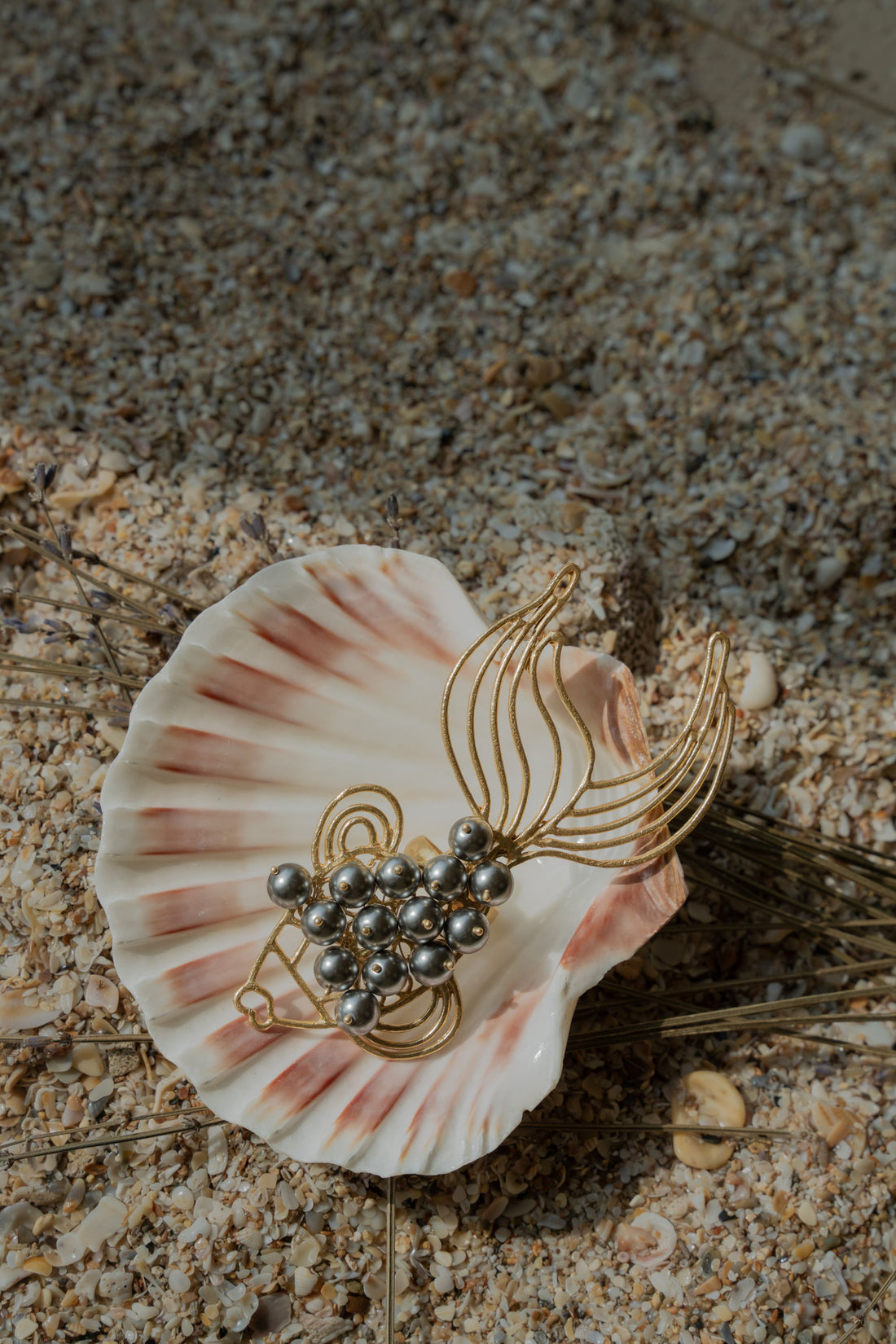 Gold-plated fish-shaped statement ring with dark grey pearl accents displayed inside a seashell on sand.