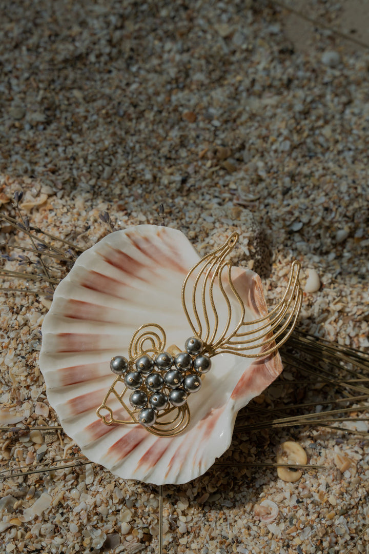 Gold-plated fish-shaped statement ring with dark grey pearl accents displayed inside a seashell on sand.