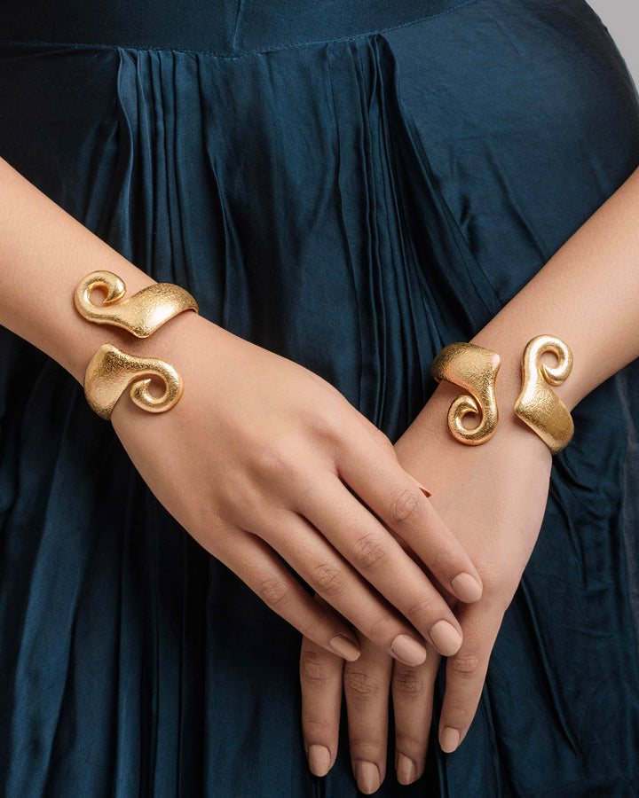 A pair of thick, textured matte-gold open cuff bracelets with a dramatic sculptural swirl design, worn on both wrists over a dark blue pleated dress.