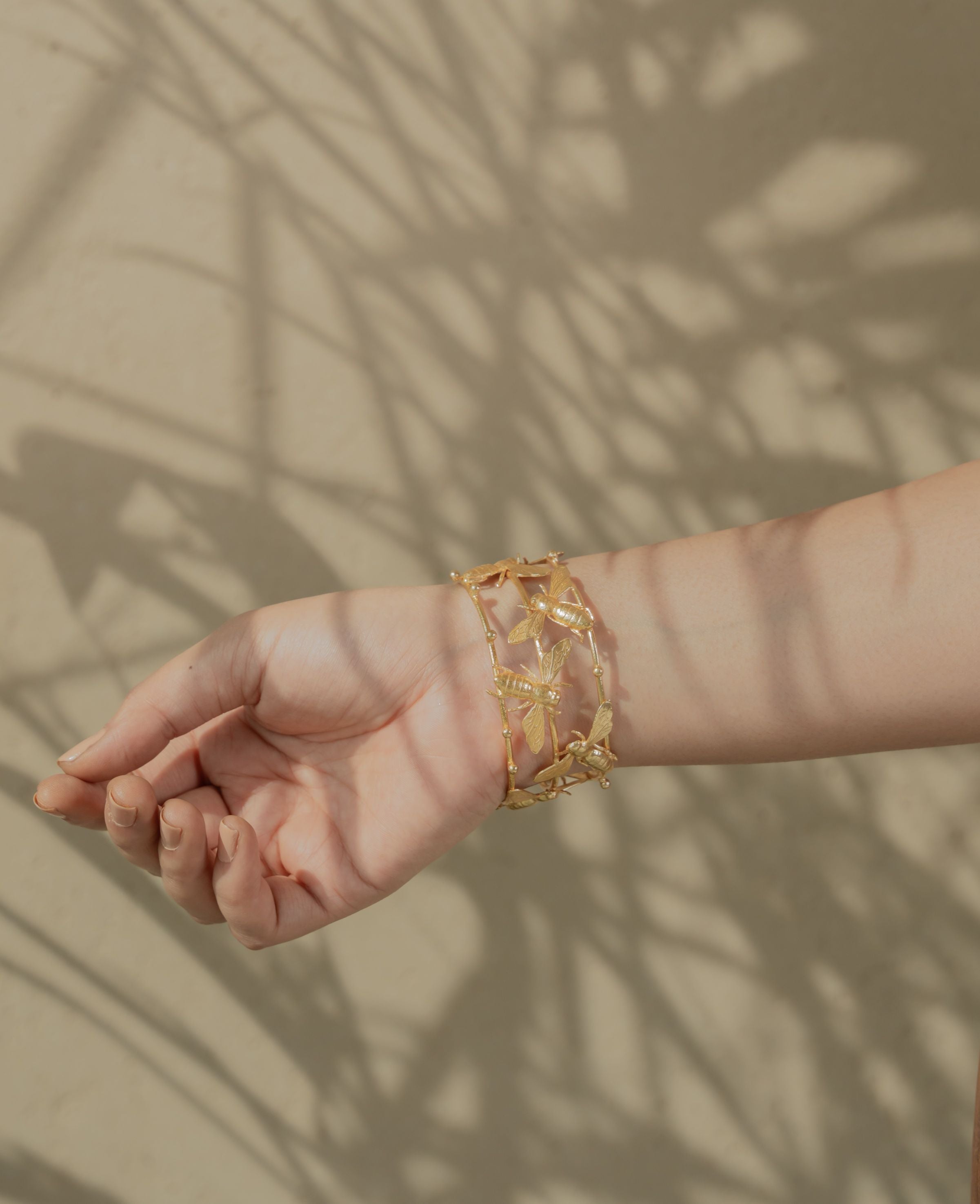 Adjustable gold-colored open cuff bracelet with bee accents resting outdoors on natural rock.