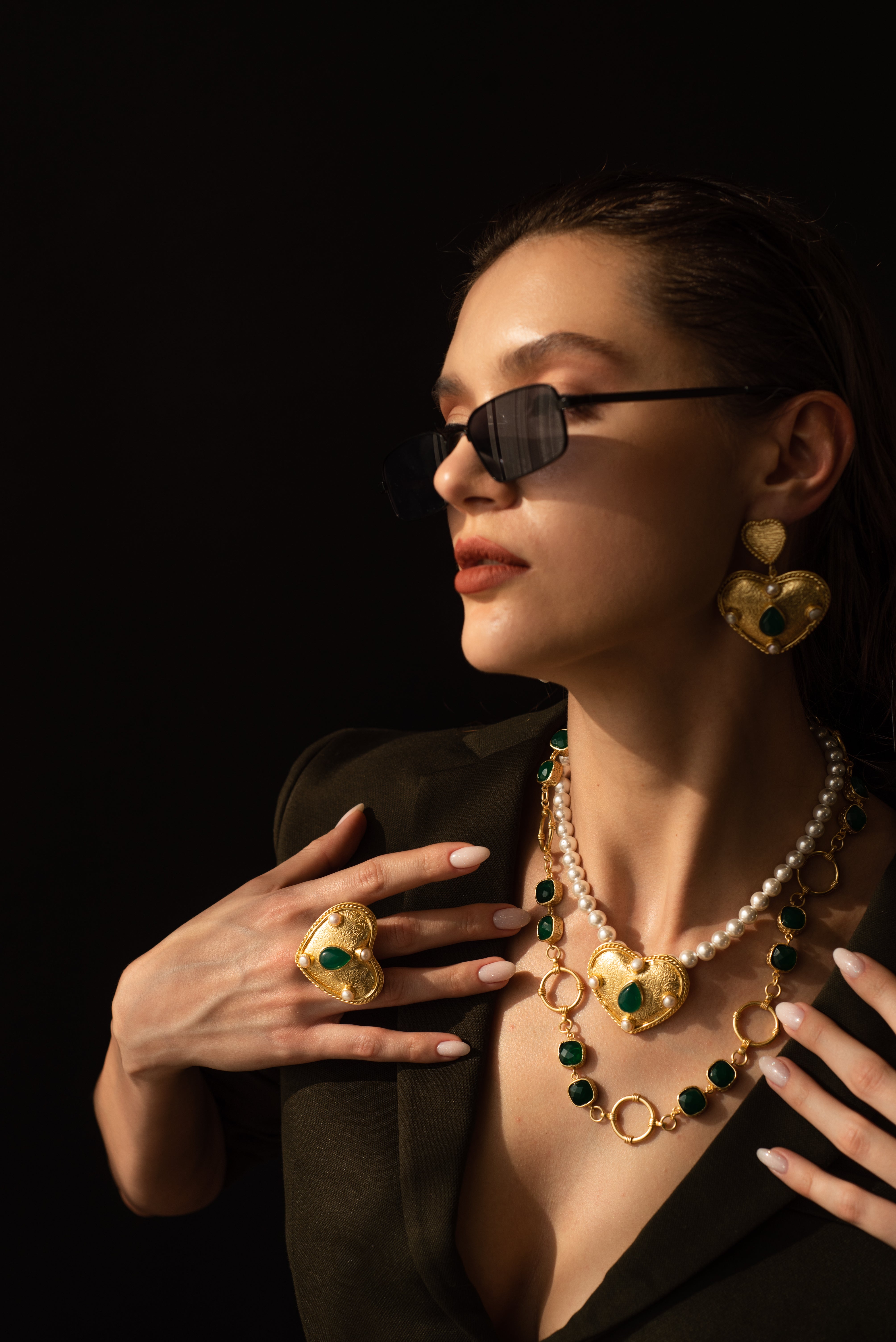 Close-up of a women's gold heart necklace featuring a green teardrop emerald center stone and pearl strand.