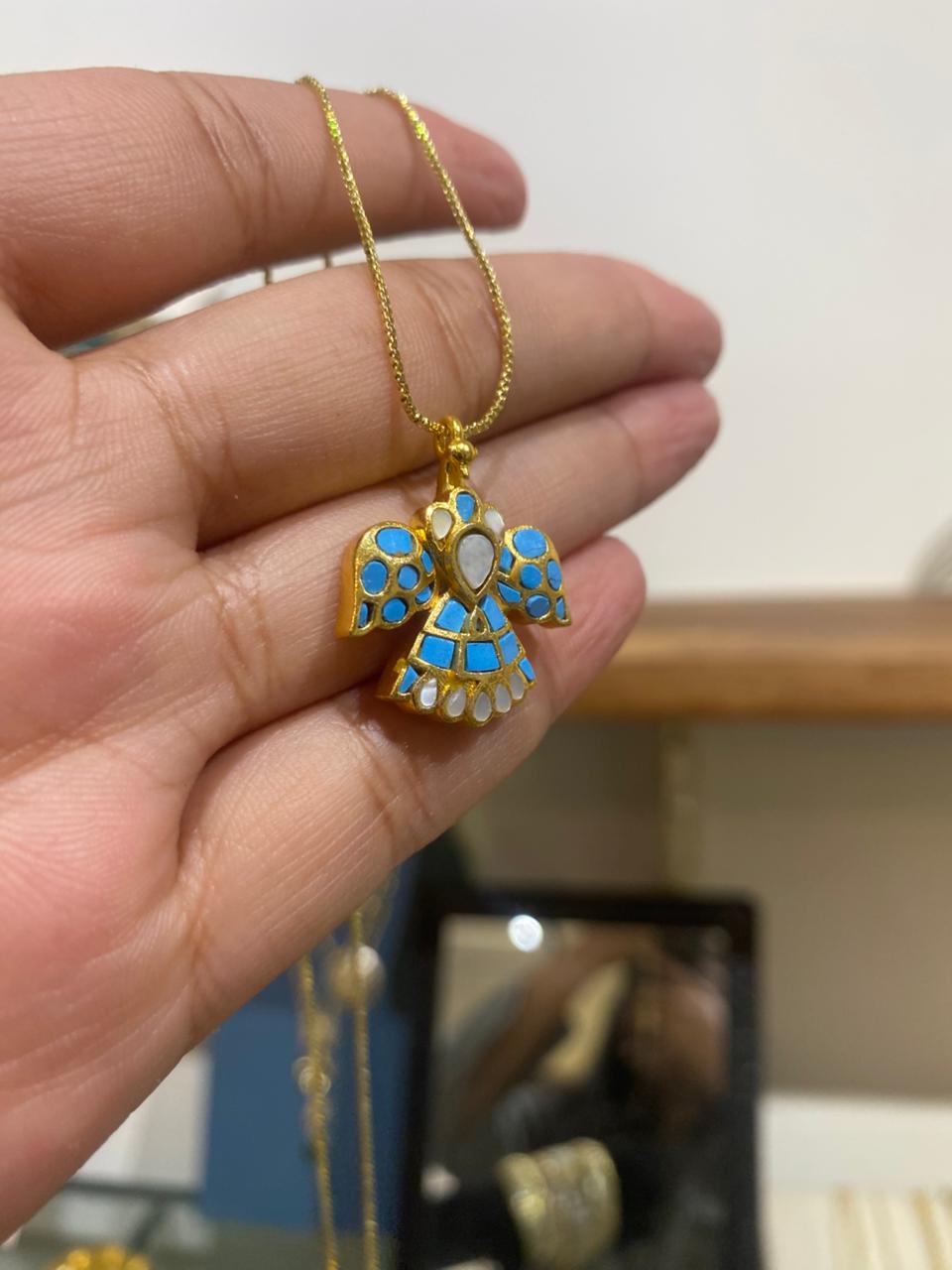 A handcrafted gold eagle pendant with vibrant turquoise and white inlay detailing, held up on a delicate gold chain.