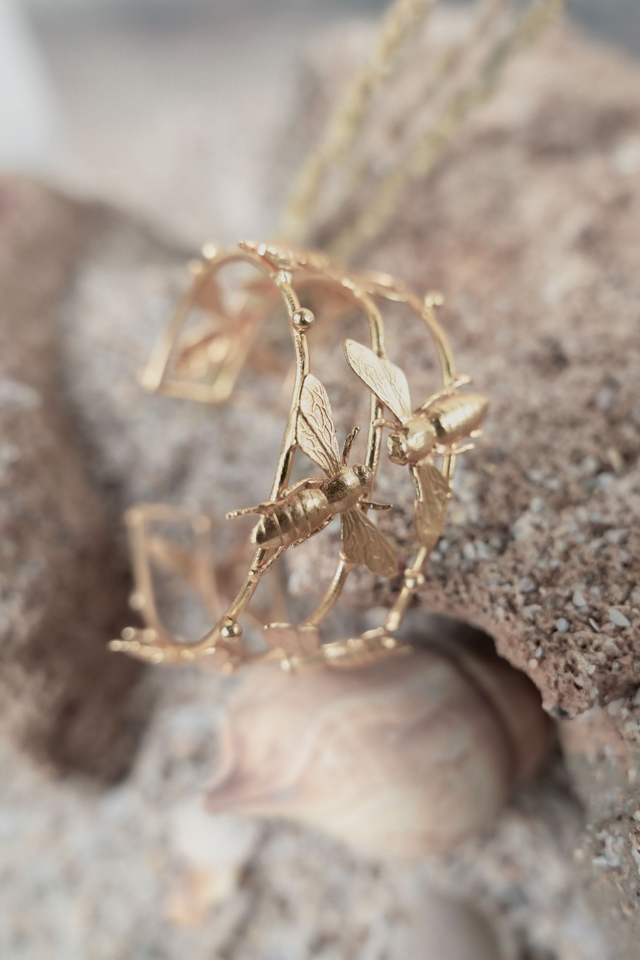 Openwork gold bangle adorned with delicate 3D bee motifs displayed on a sandy rock background.