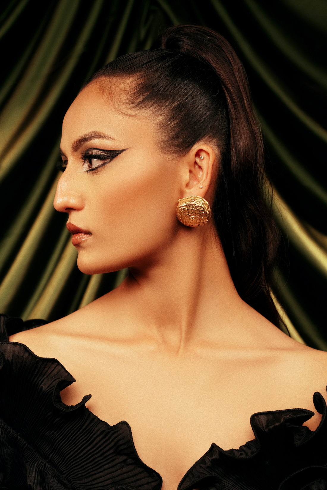 Close-up of a model wearing chunky gold Sunlit Sphere stud earrings against a dark green backdrop.