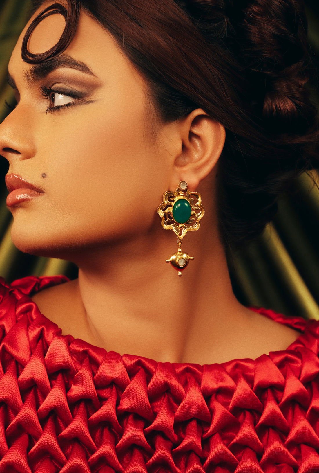 Woman styled in a textured red outfit wearing BaalaJewels Verdant Aura gold-plated statement drop earrings with a vibrant oval green gemstone.