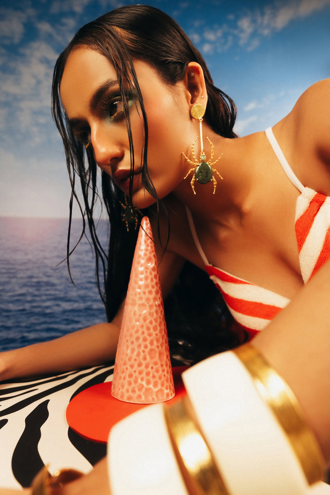 Abstract spider-web inspired gold drop earrings with dark green stones laid out on a beach-sand backdrop.