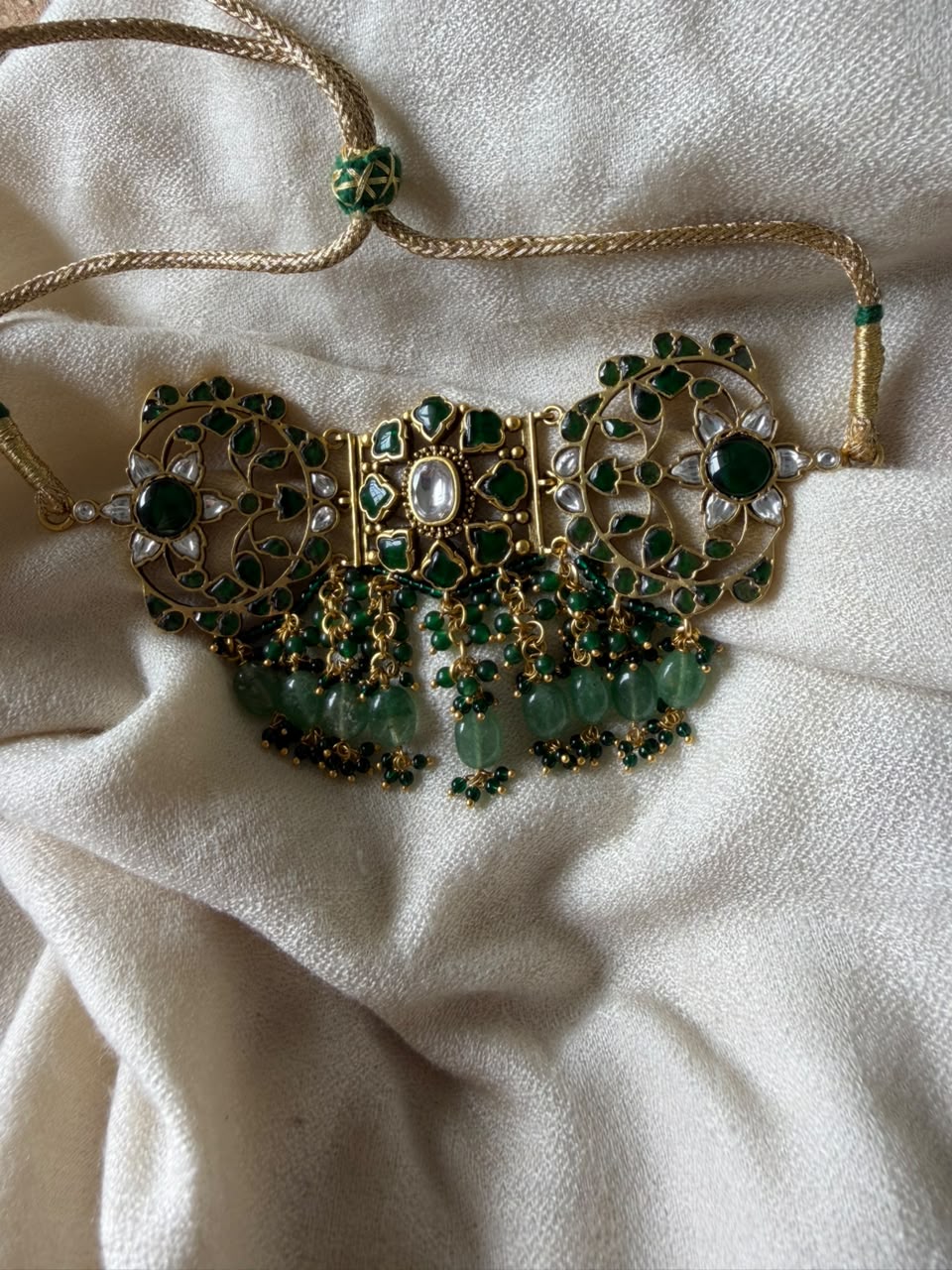 Emerald green statement Kundan choker with adjustable gold cord drawstring displayed on off-white fabric.