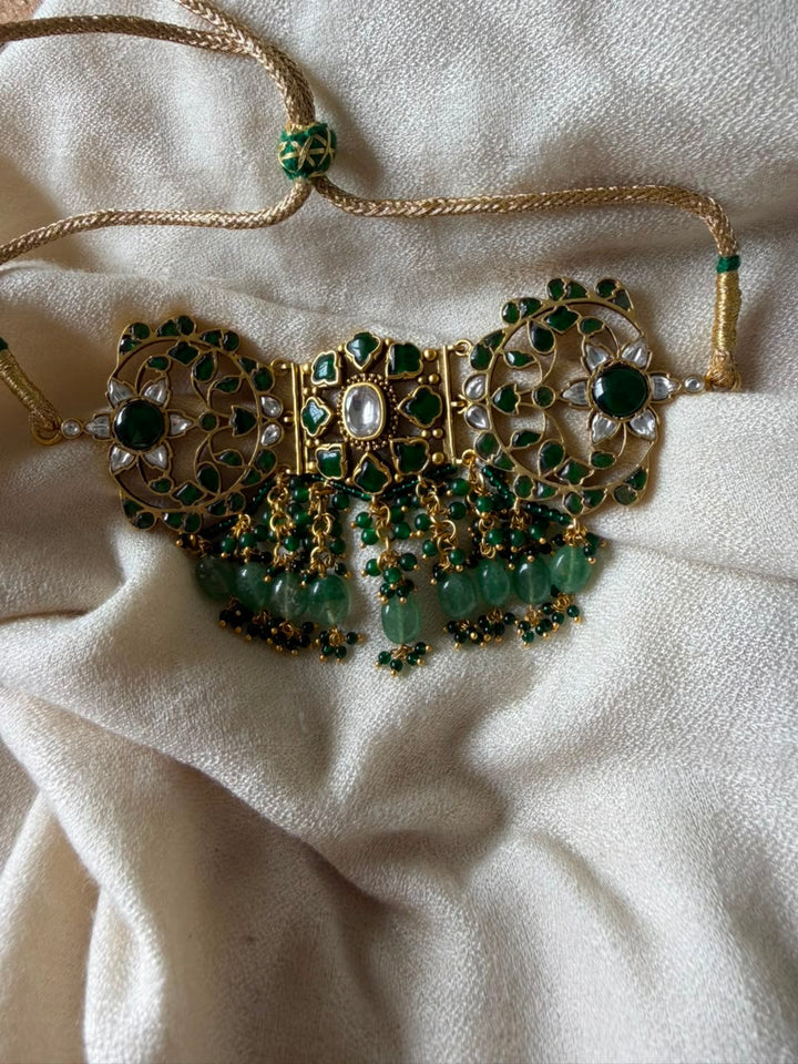 Emerald green statement Kundan choker with adjustable gold cord drawstring displayed on off-white fabric.