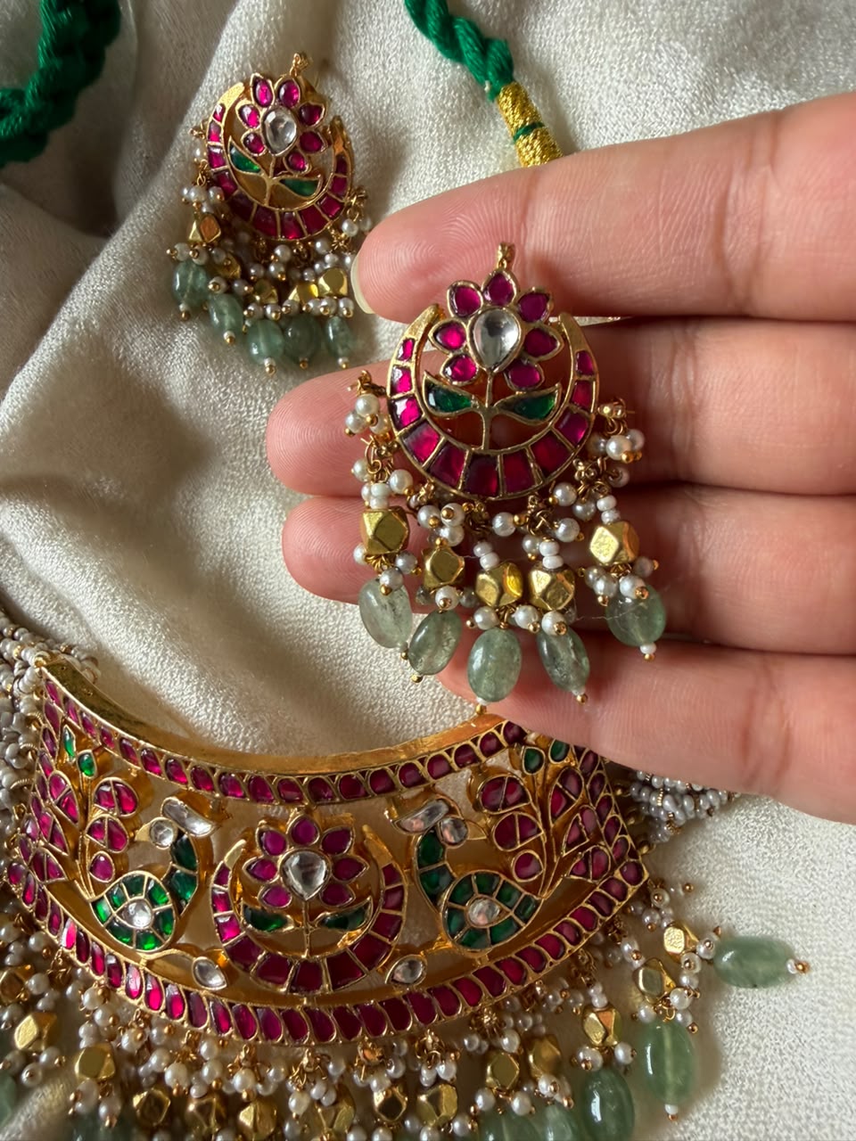 Close-up view of an intricate copper-alloy base choker necklace and matching earrings, decorated with detailed Meenakari floral artwork, precision-set Kundan glass stones, and a fringe of seed pearls with light green stones.