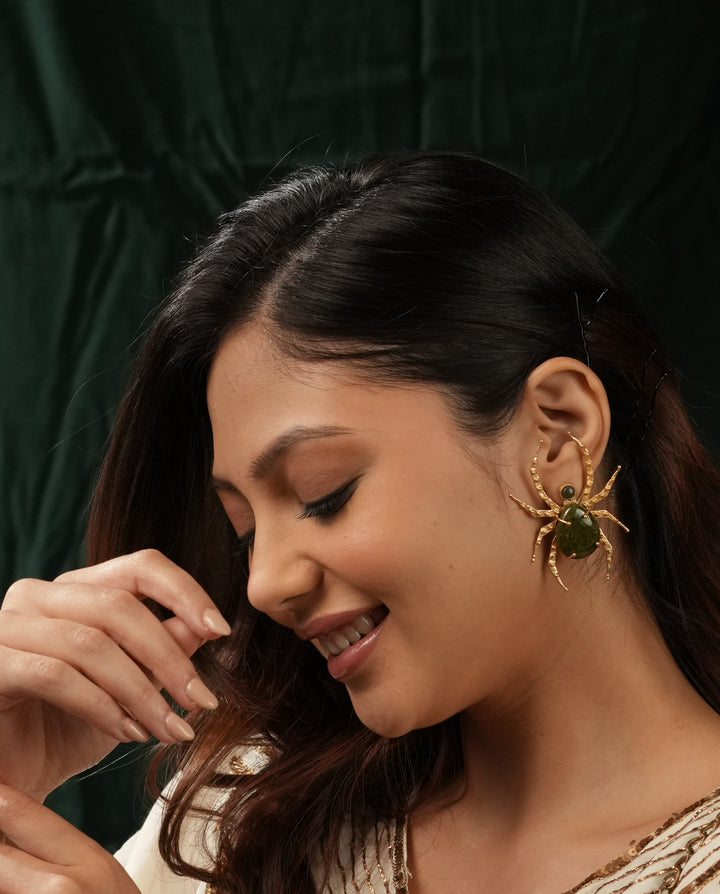 Woman wearing a gold-plated spider stud earring featuring a green gemstone body