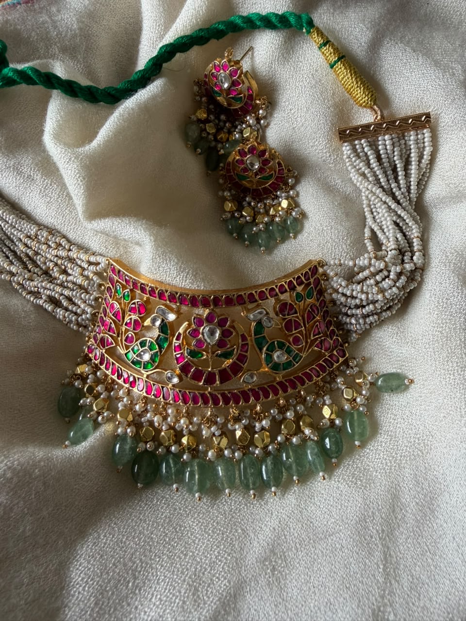 Handcrafted gold-plated bridal choker necklace and matching push-back drop earrings featuring red and green Meenakari floral motifs, white Kundan stones, multi-layered seed pearl strands, and hanging light green aventurine beads.